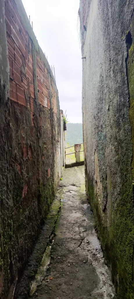 Callejón estrecho de la favela con paredes de cemento y ladrillo visto, mirando hacia la vegetación de fondo.