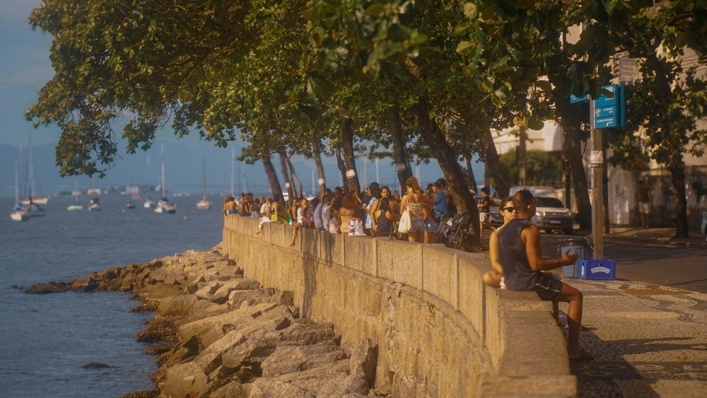 Vistas del muro de la Mureta da Urca en Río de Janeiro, con gente sentada en el borde, árboles y la bahía al atardecer.