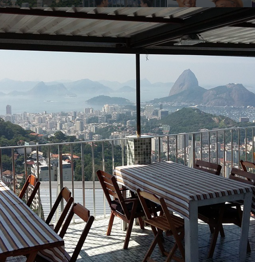 Vistas espectaculares desde la terraza techada del Bar do Tino en el Morro dos Prazeres hacia el Pão de Açúcar y la Bahía de Guanabara, Río de Janeiro, con mesas dispuestas para comer.