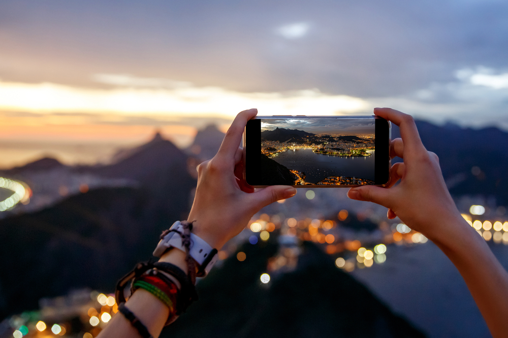 Viajera usando un smartphone para fotografiar el Pão de Açúcar al atardecer en Río de Janeiro, ilustrando la necesidad de internet en Brasil.
