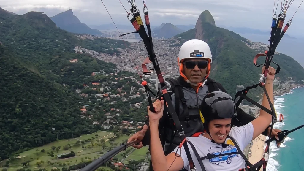 Tándem de parapente en Río de Janeiro sobrevolando la playa de São Conrado y la favela Rocinha. Dos hombres sonriendo durante la experiencia con el instructor Magrelo. El logo Río de Verdad está visible.