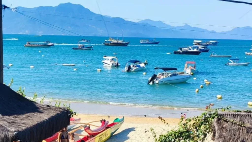 Primer plano de canoas polinesias en la arena con vista a la bahía de Vila do Abraão, Ilha Grande. El mar turquesa está lleno de barcas ancladas y montañas verdes al fondo bajo un cielo azul despejado.