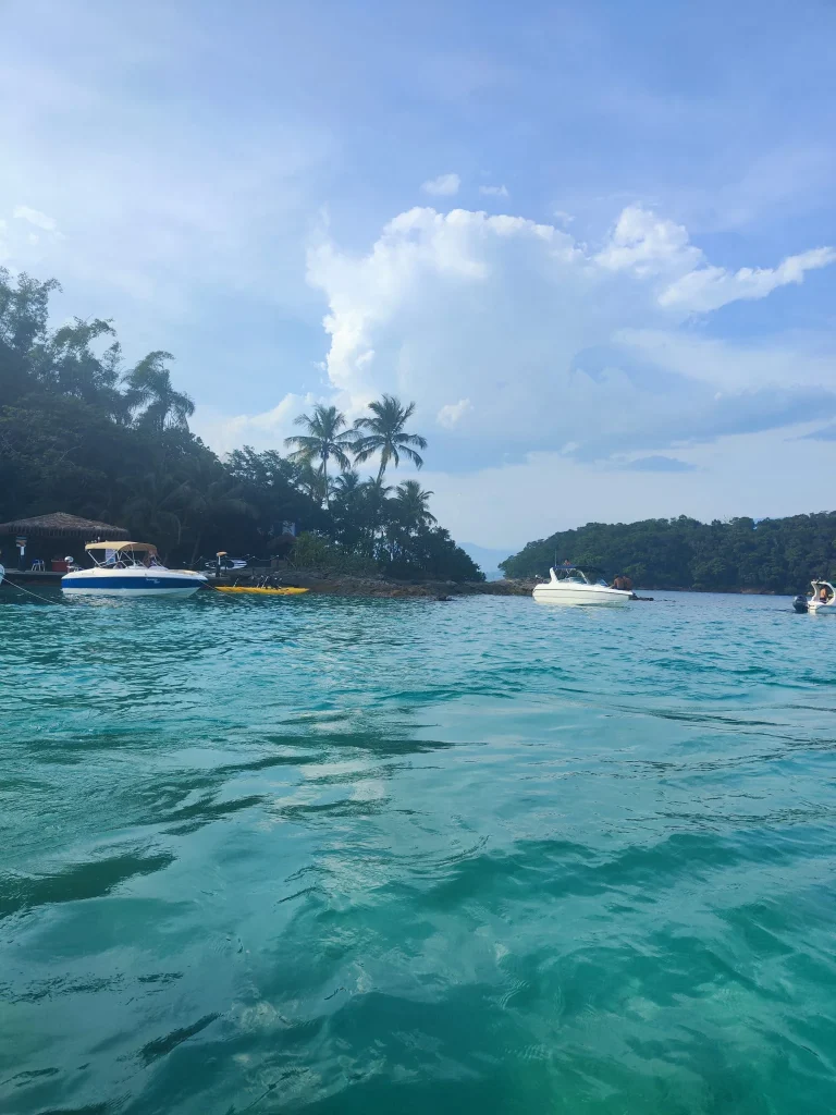 Vista desde el agua turquesa durante una parada de lancha en el tour Meia Volta en Ilha Grande. Se ven lanchas a motor ancladas cerca de una orilla con densa vegetación tropical y palmeras bajo un cielo azul con nubes blancas.