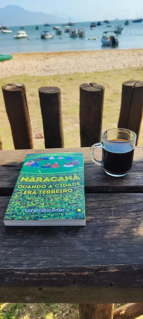 Un primer plano de un libro sobre cultura brasileña y una taza de café negro sobre una mesa de madera rústica en primera línea de playa. Al fondo, a través de troncos, se ve la bahía turquesa de Vila do Abraão con barcos de pescadores bajo un cielo despejado.