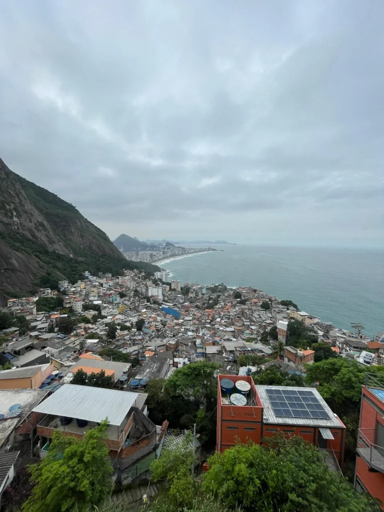 Fotografía panorámica desde lo alto del Morro Dois Irmãos mostrando la favela de Vidigal extendiéndose hasta el mar, con las playas de Ipanema y Leblon al fondo bajo un cielo nublado.
