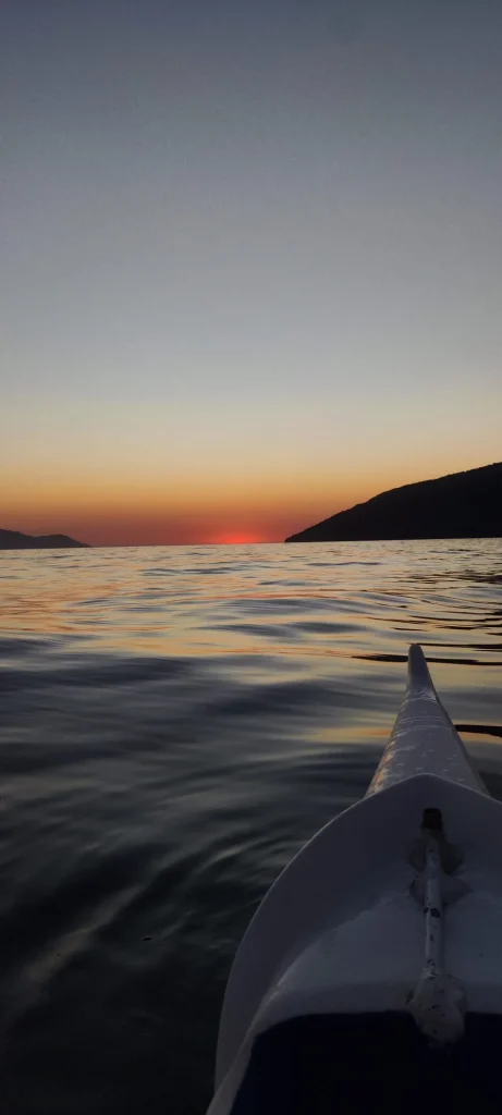 Vista desde la proa de una canoa polinesia durante el amanecer en el mar de Ilha Grande, con el sol saliendo tras las montañas de Angra dos Reis en un cielo naranja.