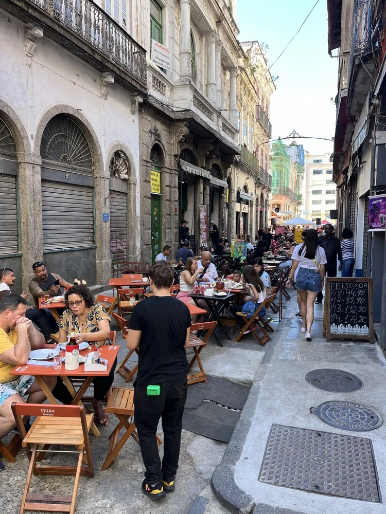 Mesas y sillas de madera en la calle peatonal adoquinada Rua do Ouvidor en el Centro de Río de Janeiro, con gente almorzando frente a bares tradicionales y un cartel que anuncia feijoada.