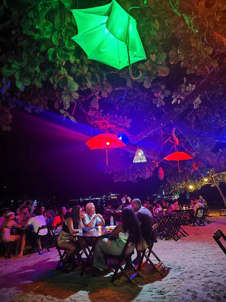Restaurante nocturno en la playa de Vila do Abraão, Ilha Grande, con mesas sobre la arena y decoración de paraguas iluminados en rojo y verde bajo los árboles.