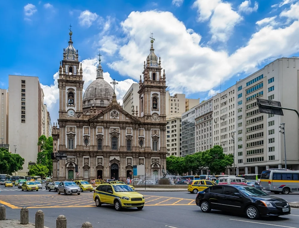 Vista frontal de la imponente Iglesia de Nuestra Señora de la Candelária con sus torres neoclásicas, ubicada en una concurrida intersección del Centro de Río de Janeiro con taxis amarillos y tráfico.