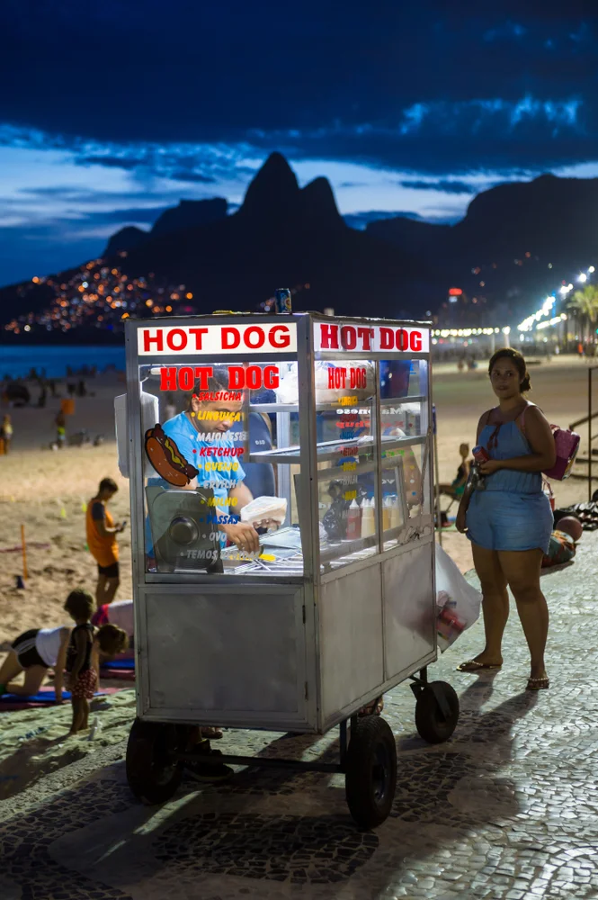 Preparación de un cachorro-quente brasileño en un carrito callejero. Se ve el pan abierto con salchicha, gran cantidad de batata palha (patatas paja), maíz y chorros generosos de mostaza y mayonesa casera antes de ser prensado.