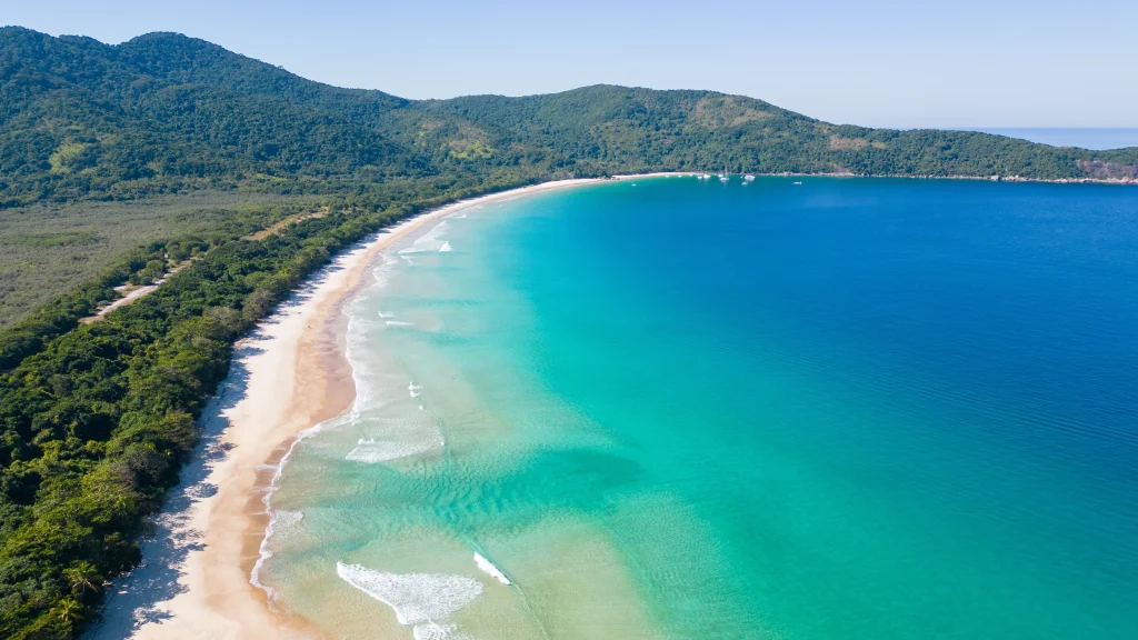 Vista aérea de la playa de Lopes Mendes en Ilha Grande. Se ve una larga franja de arena blanca y virgen, aguas turquesas y azules cristalinas con pequeñas olas, y una densa vegetación de mata atlántica verde esmeralda rodeando la bahía bajo un cielo azul despejado.
